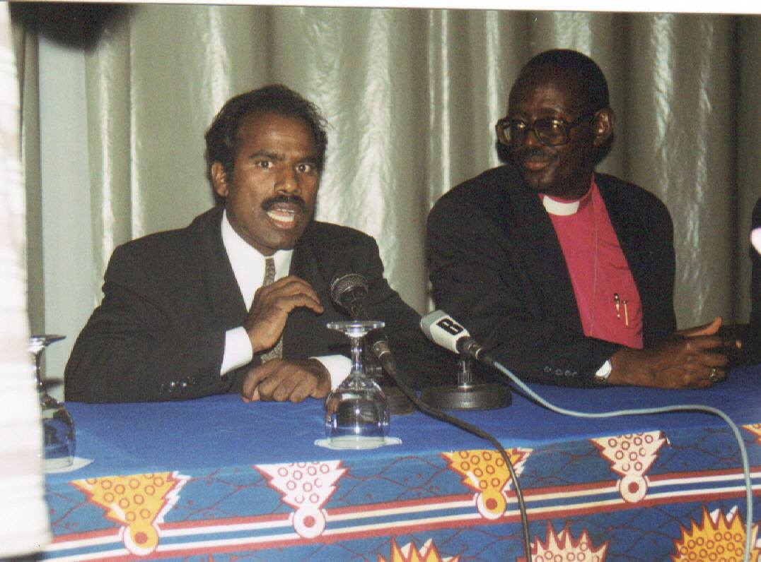 Dr. K.A. Paul speaking at a press table alongside a bishop